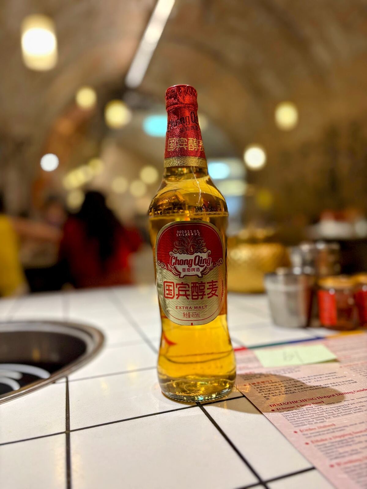 A bottle of Chong Qing Extra Malt beer on the tiled hotpot table, with the brick arches of the air-raid shelter behind