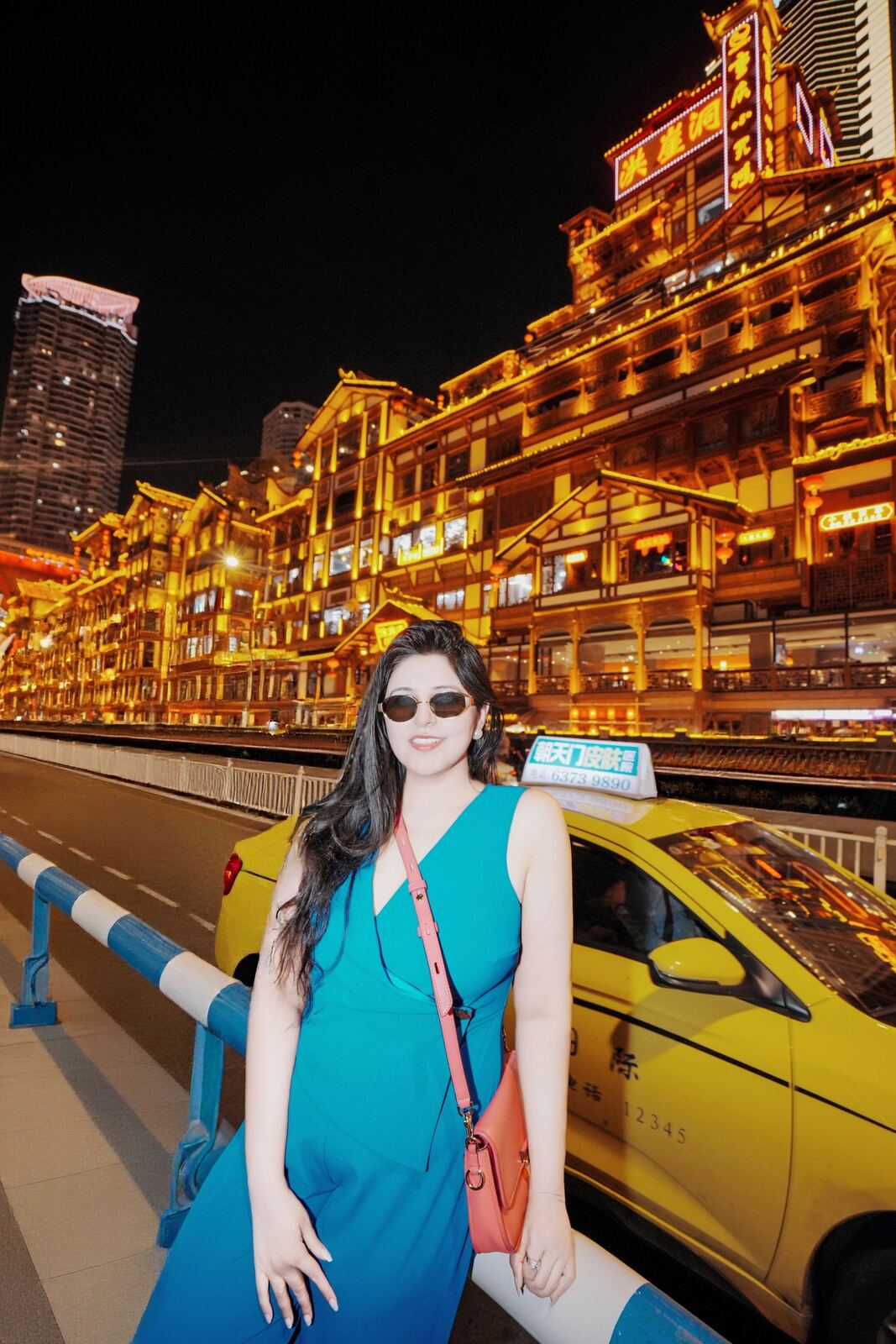 Reema in a teal dress in front of the lit-up Hongya Cave buildings at night in Chongqing, with a yellow taxi behind her