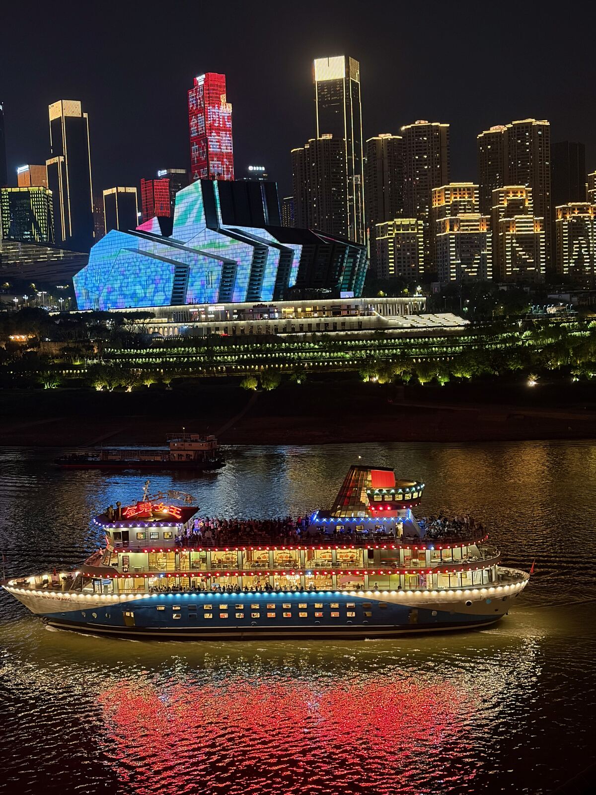 A lit-up river cruise boat in front of the glowing Chongqing skyline at night