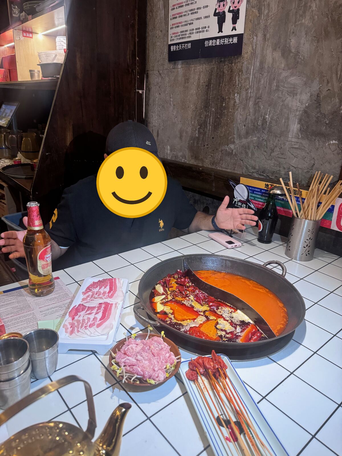 My husband at the hotpot table with a split pot, one half red with chillies and the other a milder orange broth, next to plates of sliced meat, skewers and a beer